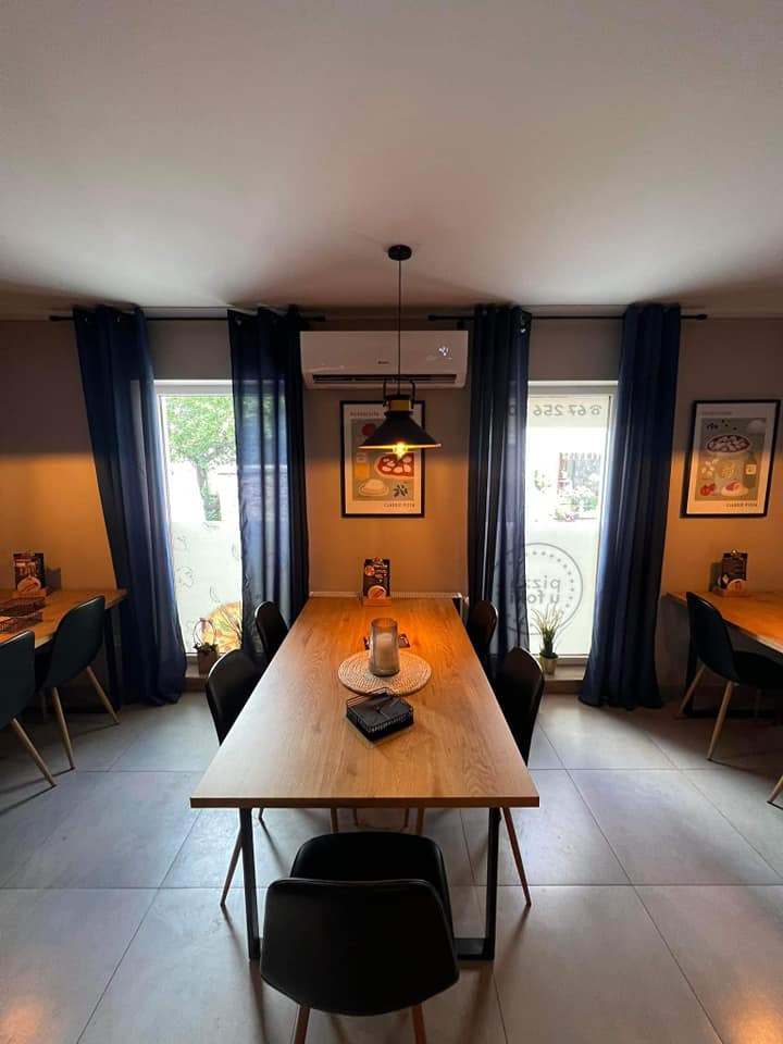 Wnętrze restauracji z długim drewnianym stołem, czarnymi krzesłami, zasłonami w oknach i zamontowanym klimatyzatorem.