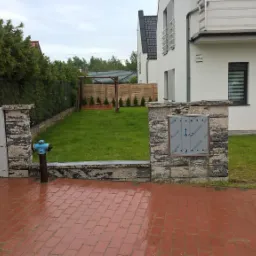 Czerwona kostka brukowa na podjeździe, ograniczona kamiennymi słupkami i skrzynkami z oznaczeniami gazu i elektryczności, w tle trawnik, huśtawka i nowoczesny dom z białymi ścianami.