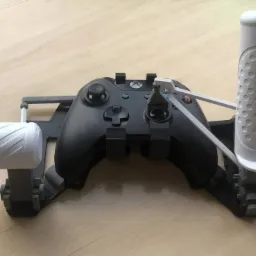 Czarny kontroler Xbox z przymocowanym, drukowanym w 3D, szarym uchwytem z białymi cylindrycznymi elementami po bokach, leżący na drewnianym blacie.