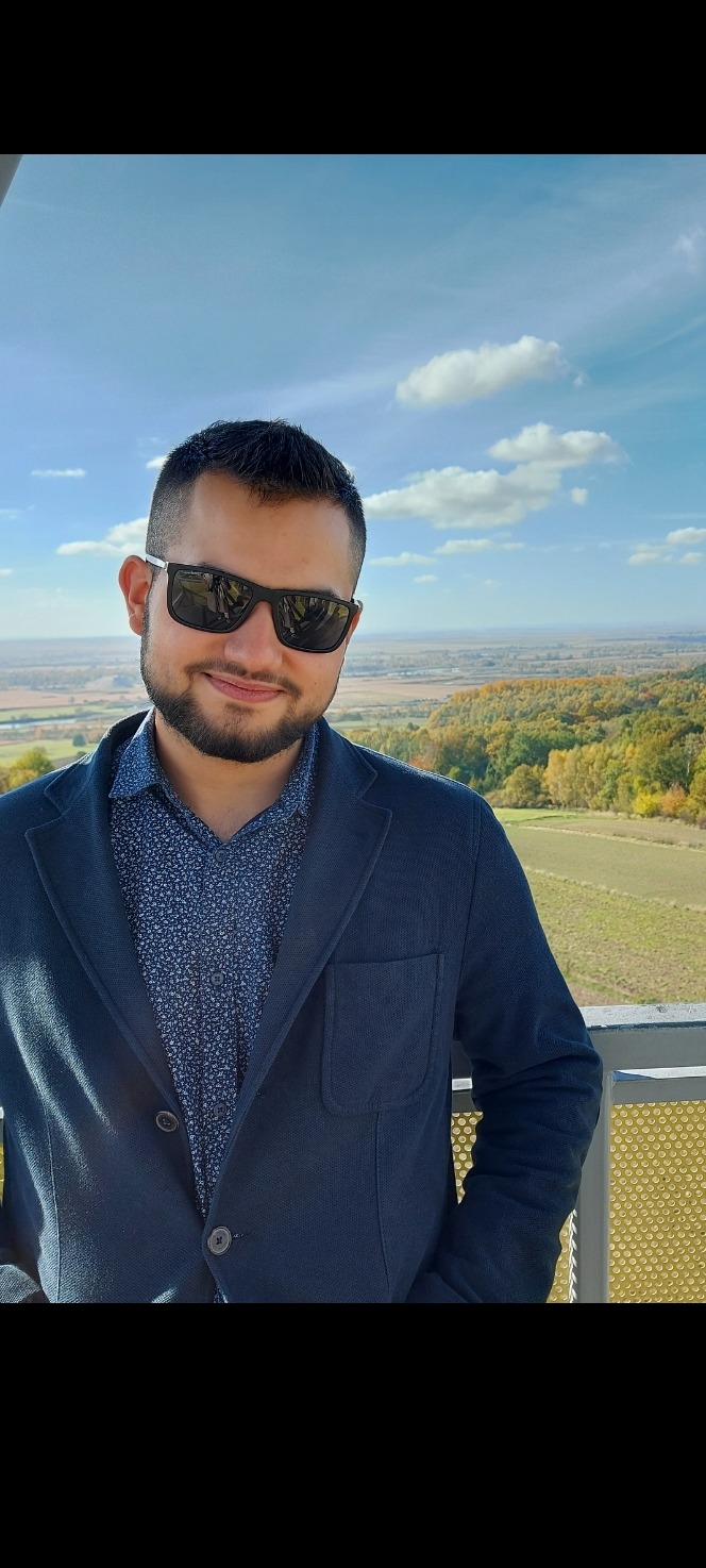 Portret mężczyzny w okularach przeciwsłonecznych i marynarce na tle jesiennego krajobrazu z błękitnym niebem i chmurami.