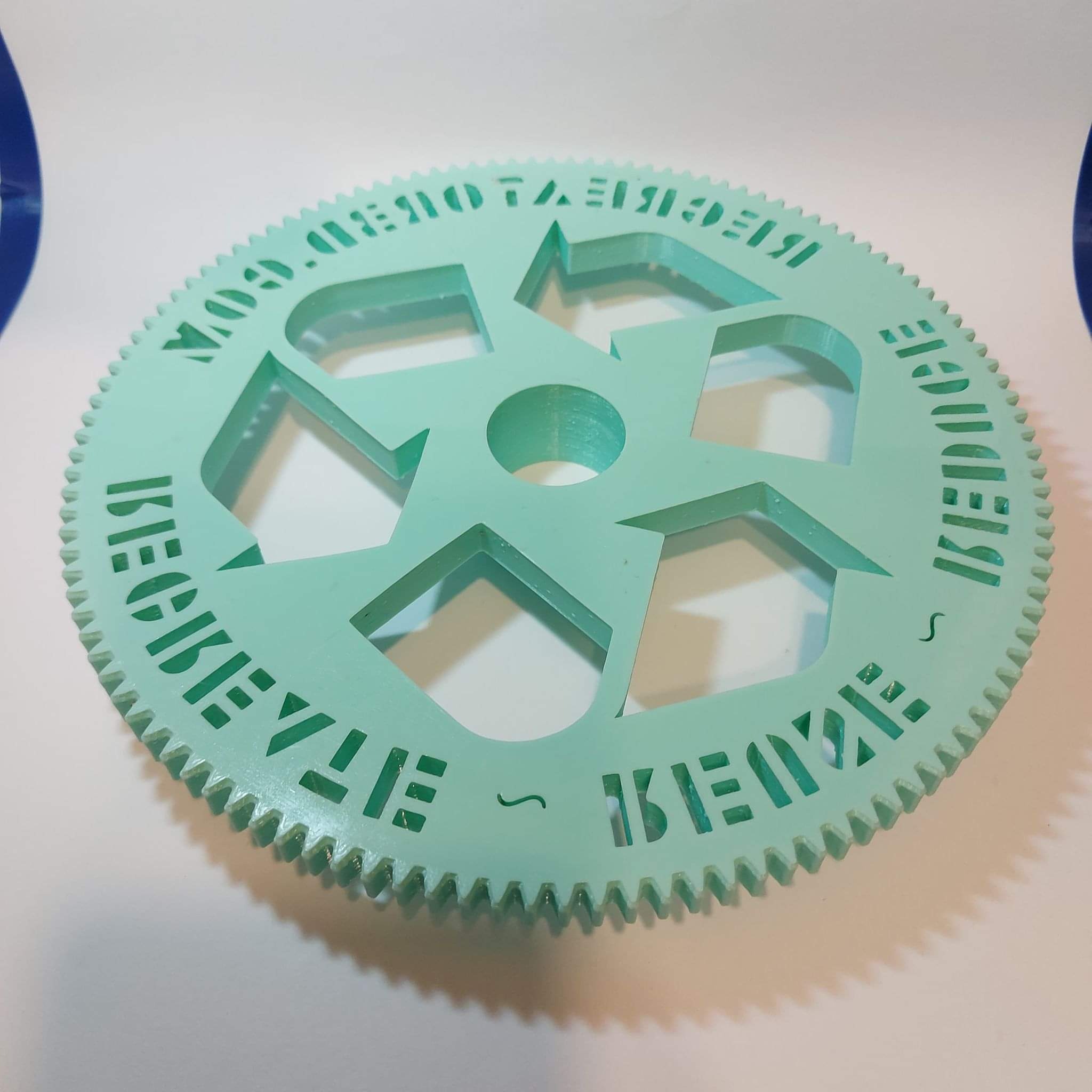 Miętowa tarcza z zębatym obwodem i wyciętym symbolem recyklingu oraz napisem 'REDUCE REUSE RECYCLE' wykonana techniką druku 3D.