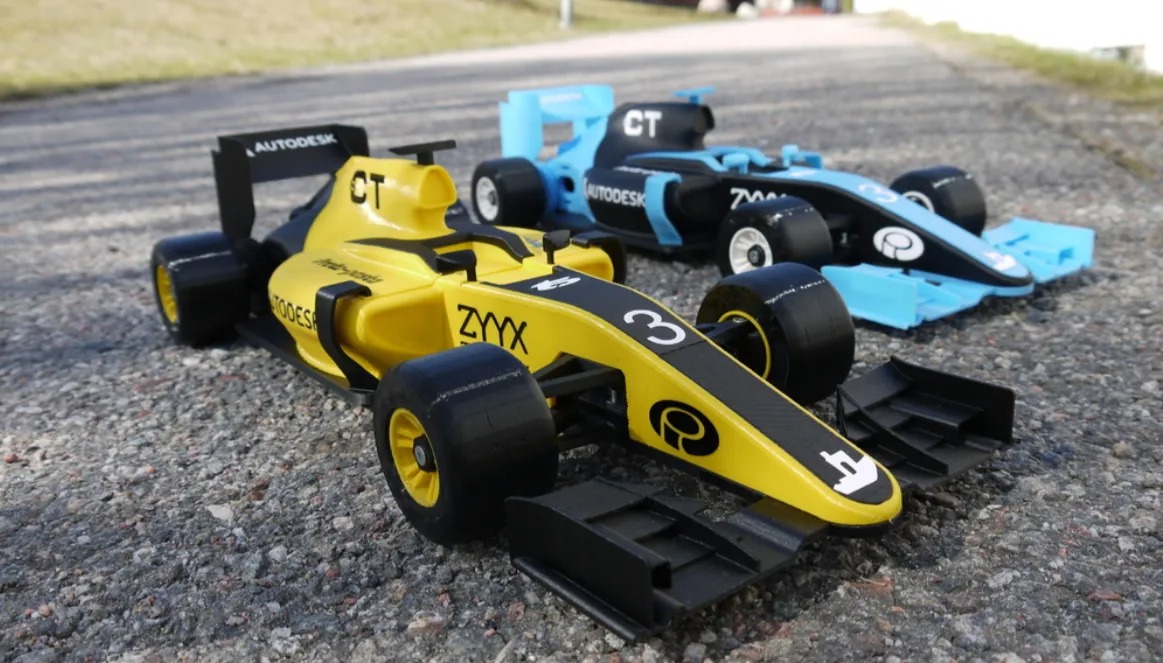 Dwa modele bolidów Formuły 1: żółty z logo ZYYX na pierwszym planie i niebiesko-czarny w tle, oba z logotypami Autodesk, ustawione na szarej, szorstkiej powierzchni.