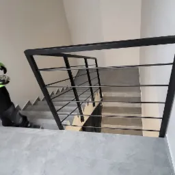 Nowoczesne schody z szarymi stopniami i czarną, minimalistyczną poręczą, widok z góry na dół, fragment sylwetki osoby w odzieży roboczej.