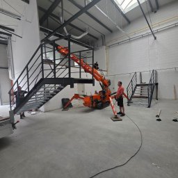 Dariusz Pasiciel - Montaż metalowych schodów w hali przemysłowej przy użyciu miniżurawia. Widoczni pracownicy i elementy konstrukcyjne schodów.