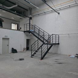 Dariusz Pasiciel - Metalowe schody w surowym wnętrzu hali produkcyjnej, widoczne instalacje pod sufitem i białe ściany z pustaków. Schody dwubiegowe z czarną balustradą.