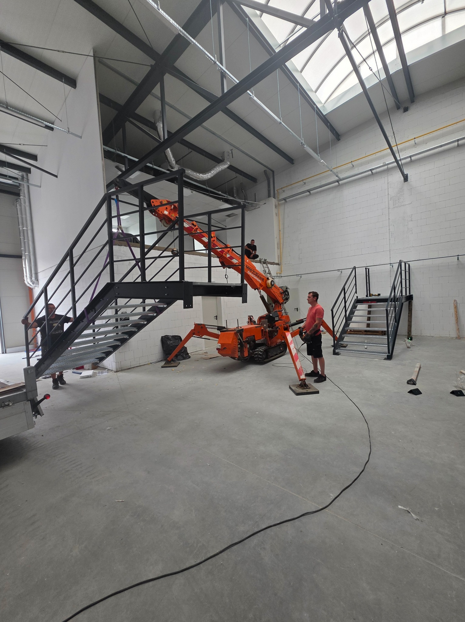 Montaż metalowych schodów w hali przemysłowej przy użyciu miniżurawia. Widoczni pracownicy i elementy konstrukcyjne schodów.