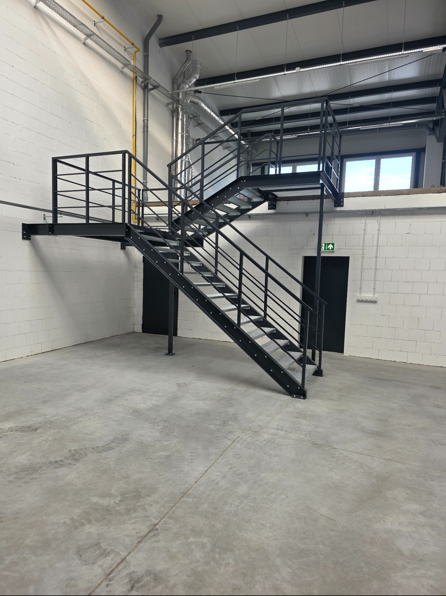 Czarne schody metalowe z platformą w industrialnym wnętrzu, widok z dołu. Białe ściany z bloków, betonowa podłoga, rury pod sufitem. Widoczny znak ewakuacyjny.
