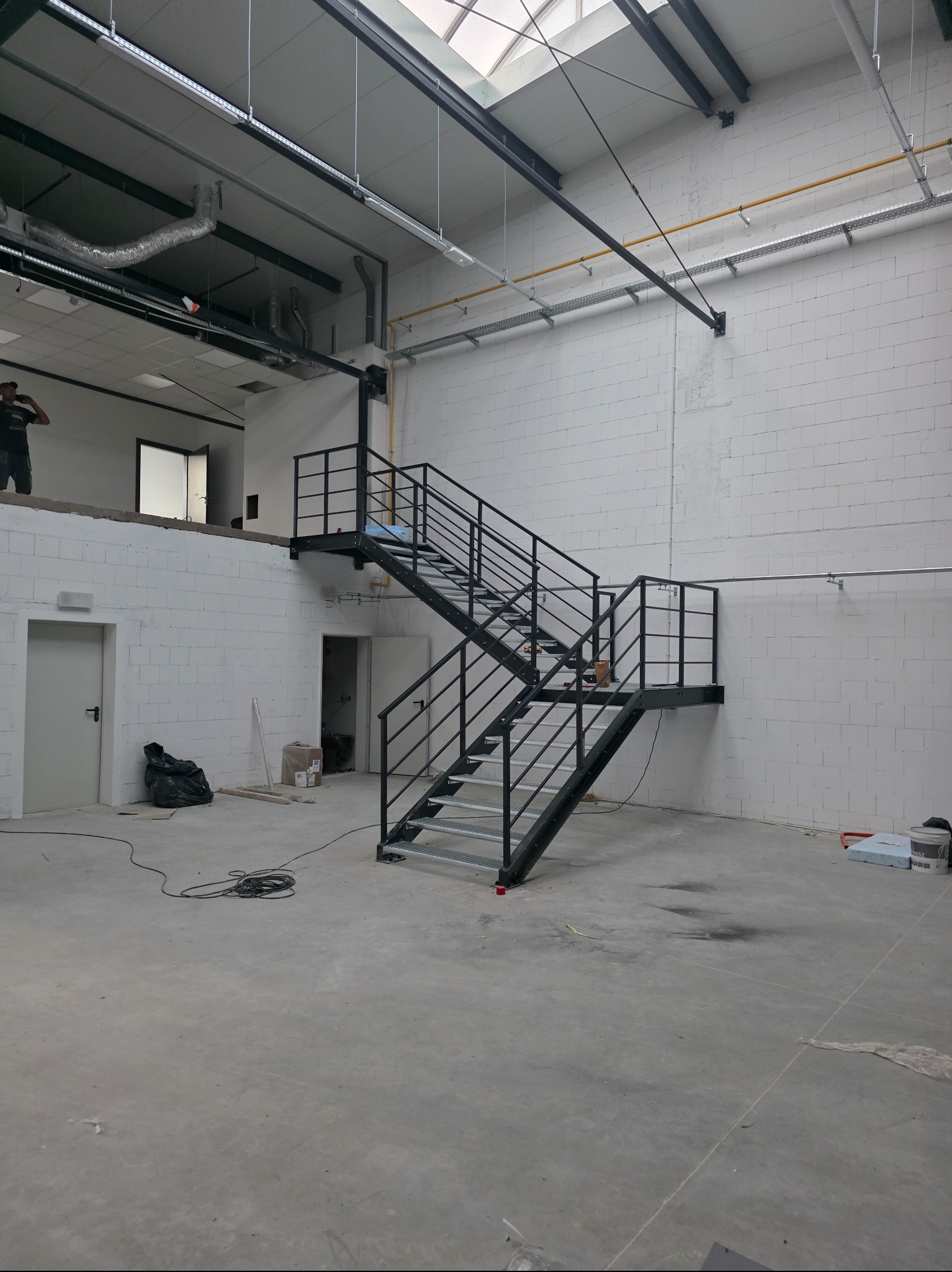 Metalowe schody w surowym wnętrzu hali produkcyjnej, widoczne instalacje pod sufitem i białe ściany z pustaków. Schody dwubiegowe z czarną balustradą.