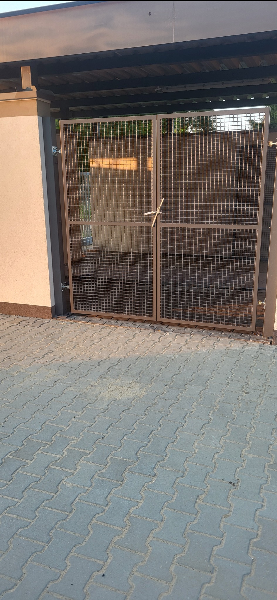 Metalowa, dwuskrzydłowa brama z siatki, z prowizorycznym zamknięciem ze sznurka, zamontowana przy budynku z kostką brukową. Widoczne zawiasy i elementy konstrukcyjne.