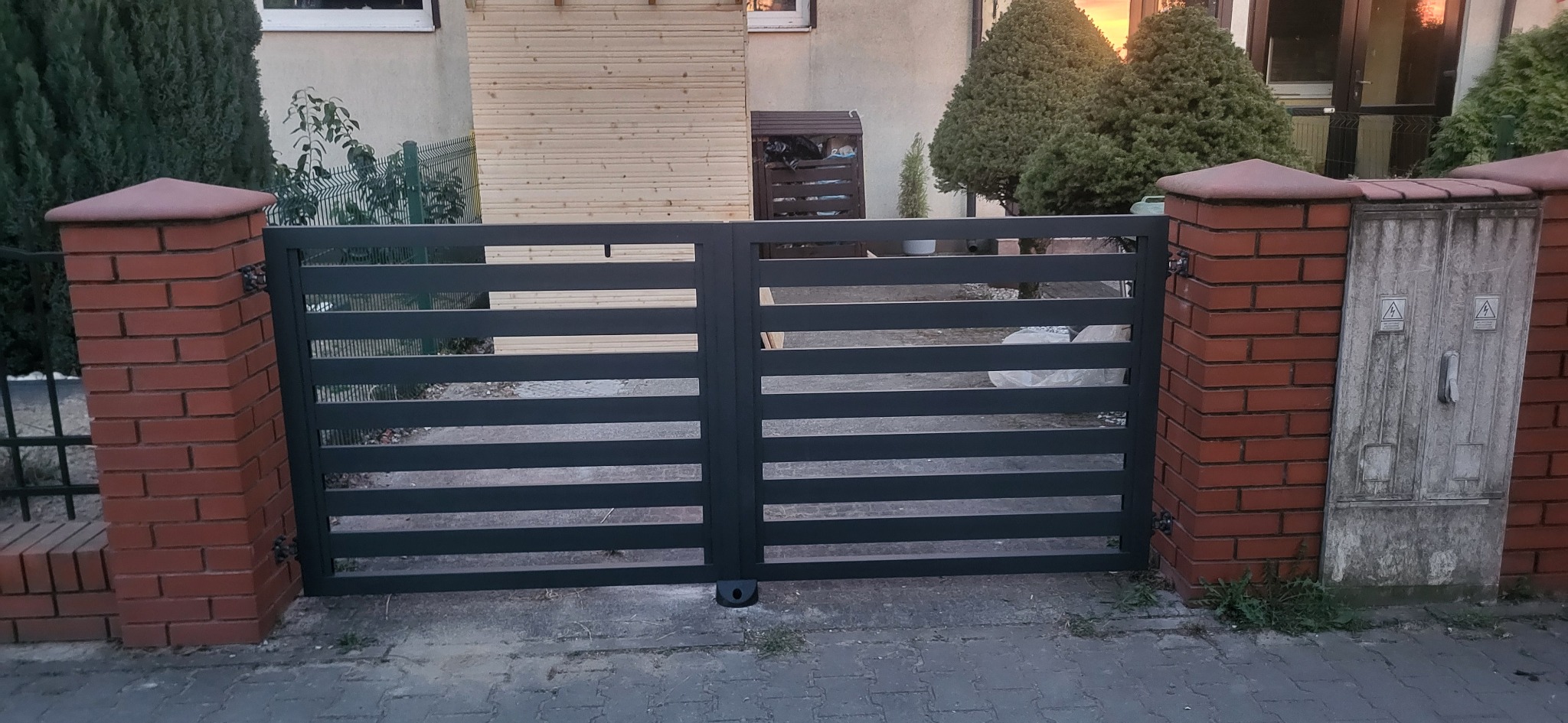 Nowoczesna, pozioma brama metalowa w kolorze grafitowym, osadzona między dwoma ceglanymi słupkami. Tło stanowi budynek i zieleń.