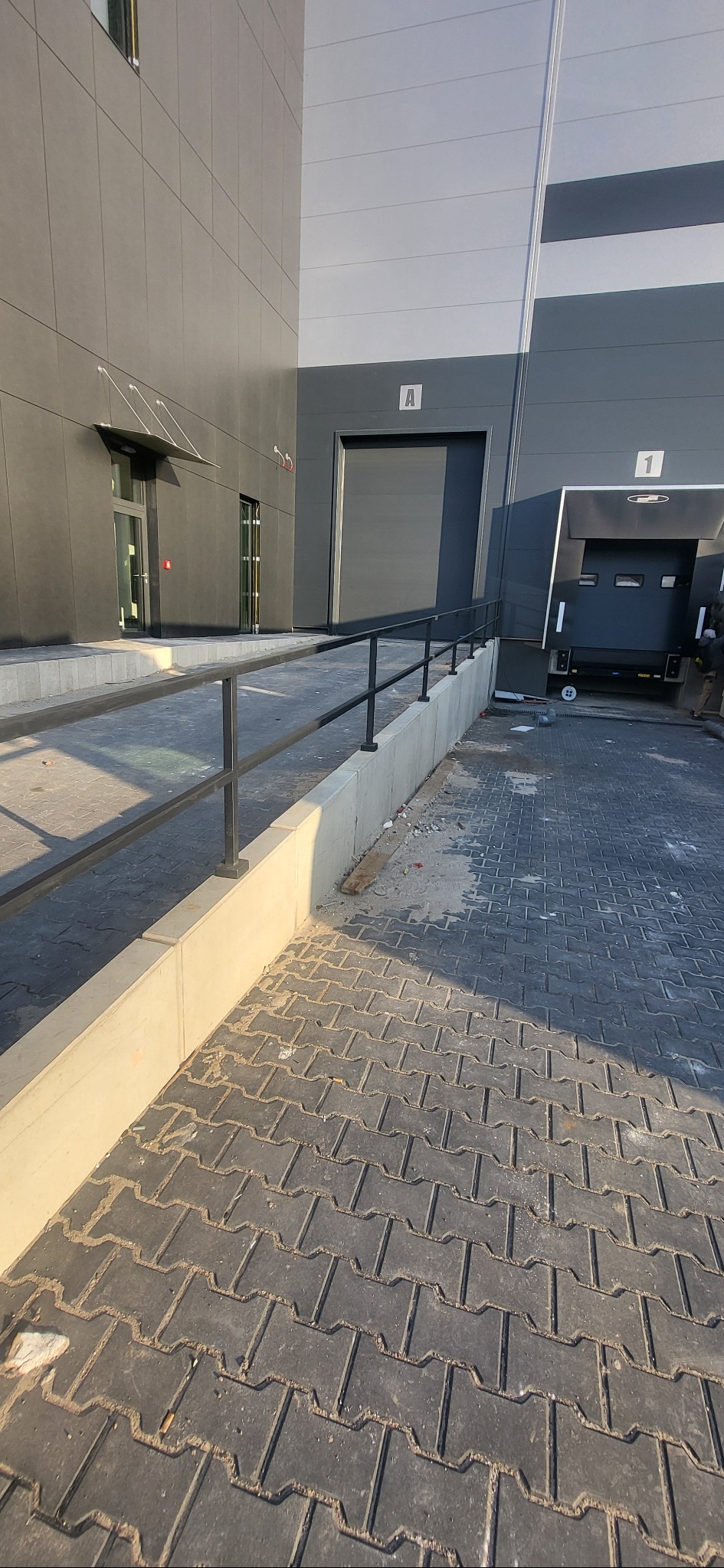 Zewnętrzna poręcz wzdłuż betonowego murku przy rampie wjazdowej do budynku magazynowego, z widocznymi drzwiami i bramą w tle, kostka brukowa na pierwszym planie.
