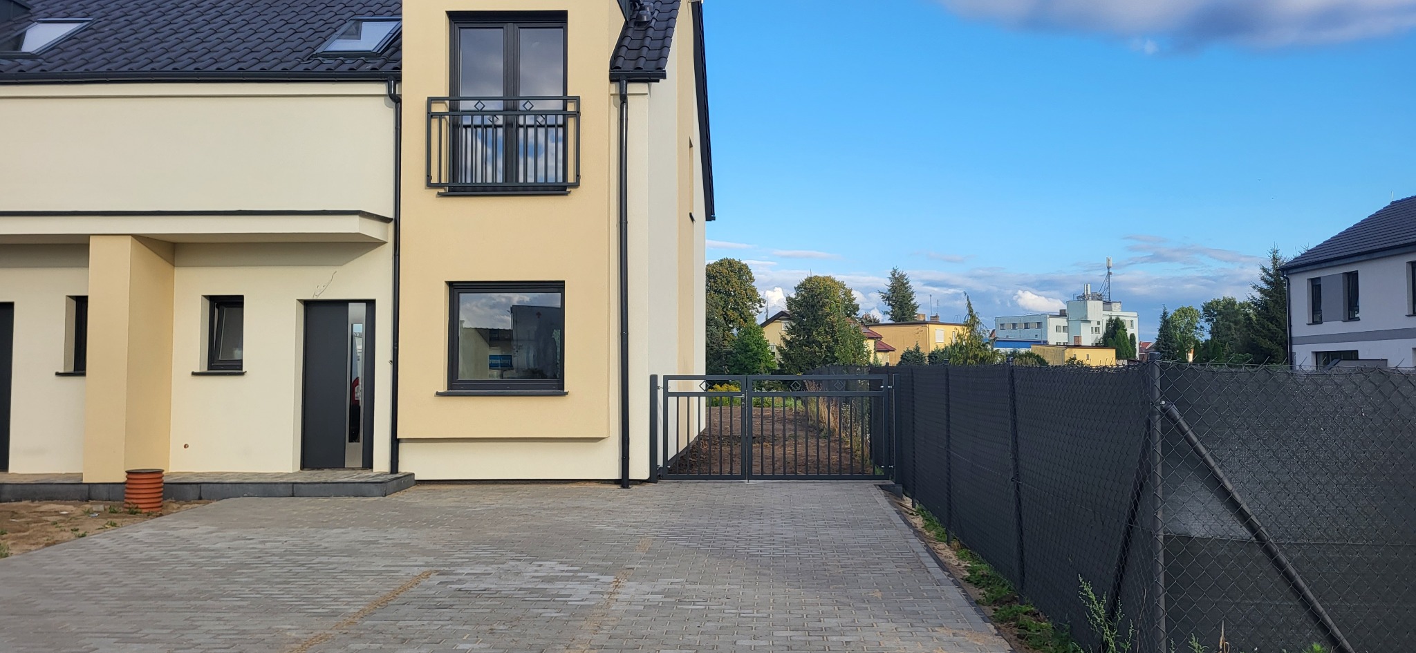 Nowoczesny dom bliźniak z czarną balustradą balkonową i bramą wjazdową, otoczony ogrodzeniem z siatki. Kostka brukowa na podjeździe, błękitne niebo w tle.