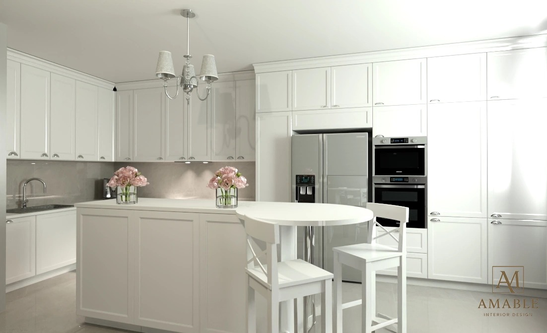 Elegancka, biała kuchnia z wyspą, okrągłym stołem i kwiatami. Klasyczne szafki, srebrny sprzęt AGD i subtelne oświetlenie tworzą spójną, jasną przestrzeń.