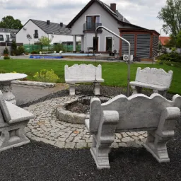 Ogród z miejscem na ognisko otoczonym betonowymi ławkami i stolikiem, w tle domy i basen.