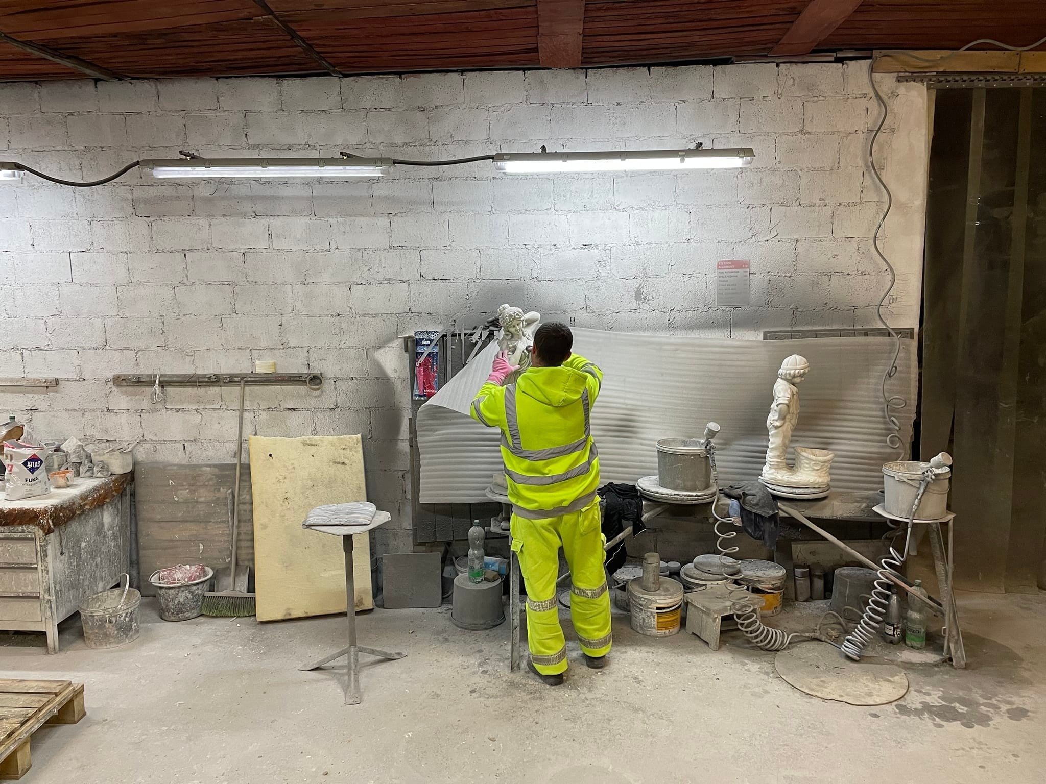 Pracownik w odblaskowym kombinezonie w warsztacie, zabezpiecza folią rzeźbę aniołka przed malowaniem natryskowym, widoczne pistolety lakiernicze i farby.