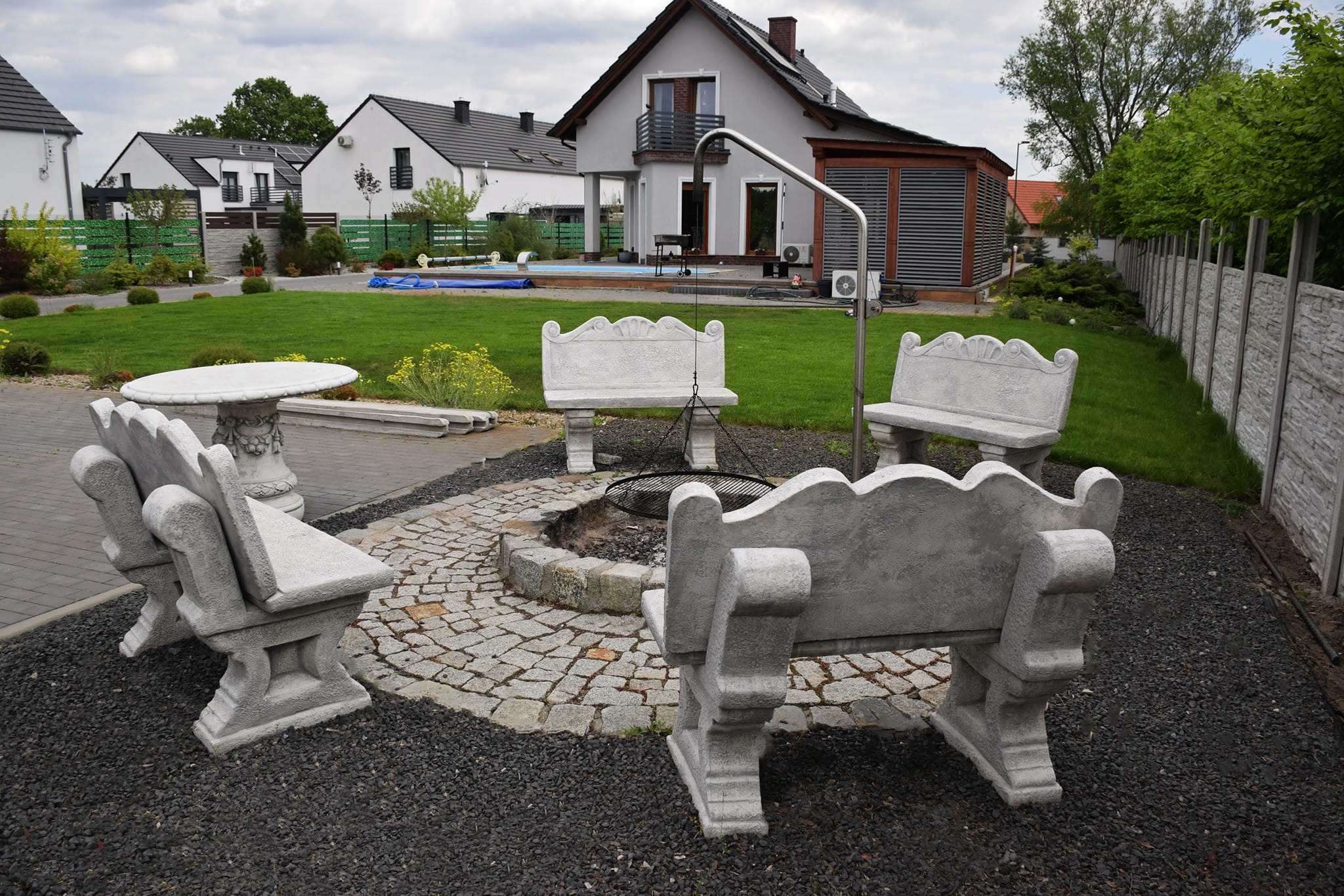 Ogród z miejscem na ognisko otoczonym betonowymi ławkami i stolikiem, w tle domy i basen.