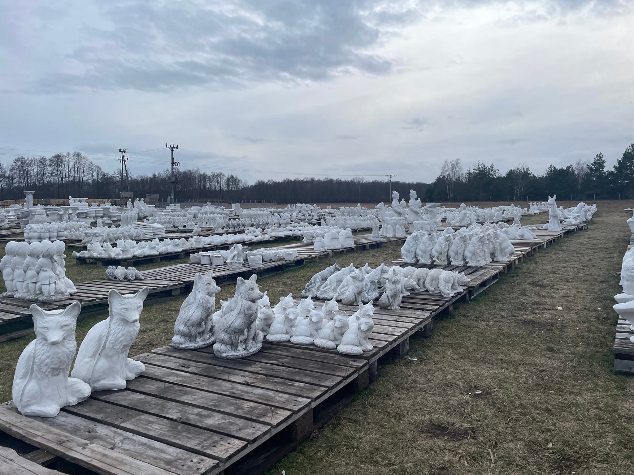 Magazyn gotowych, białych, betonowych figurek ogrodowych zwierząt i ludzi, ułożonych rzędami na drewnianych paletach, na tle pochmurnego nieba i drzew.
