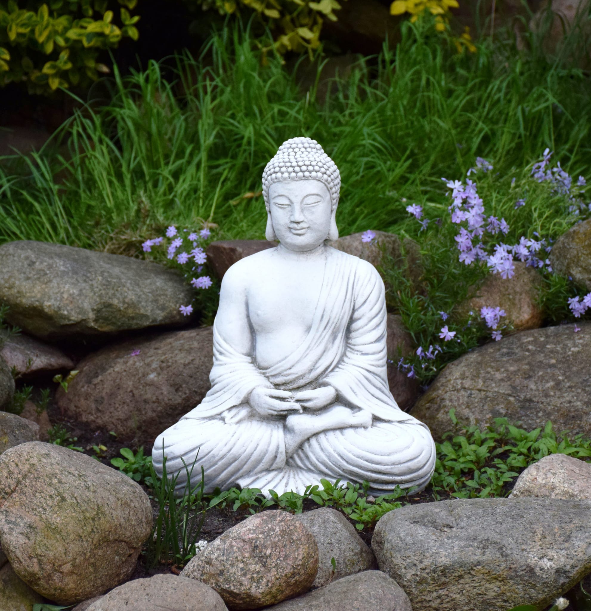 Biała statuetka Buddy w pozycji lotosu otoczona kamieniami, trawą i fioletowymi kwiatami w ogrodzie w Nowej Soli.