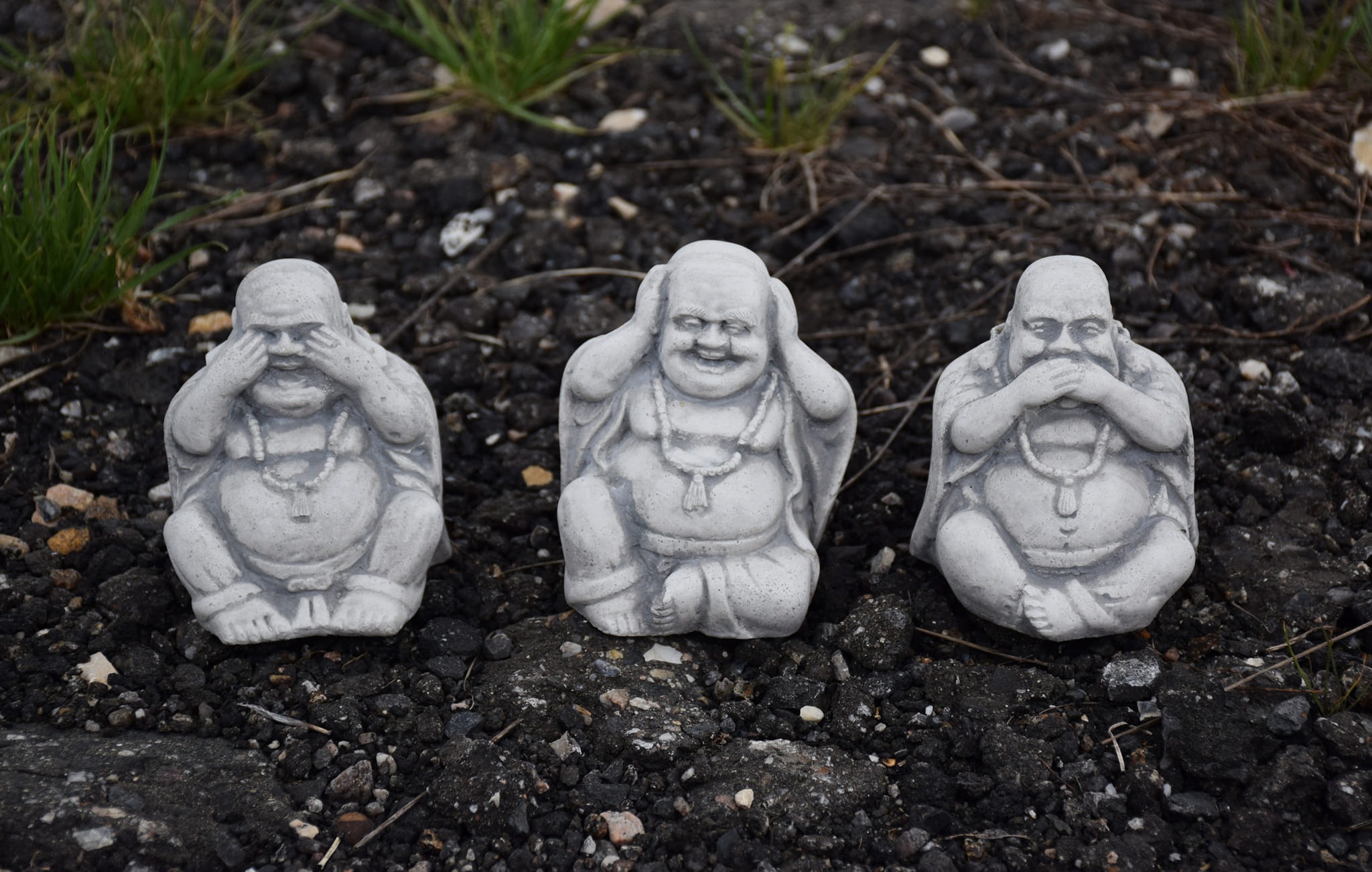 Trzy betonowe figurki Buddy (zakrywanie oczu, uszu i ust) ustawione na ciemnym, kamienistym podłożu z drobnymi źdźbłami trawy.