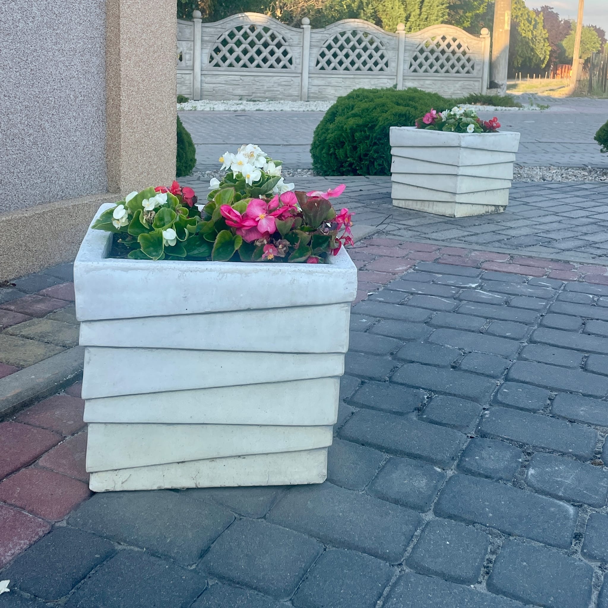 Kwadratowa, betonowa donica warstwowa z kolorowymi begoniami na brukowanym podwórku, w tle betonowe ogrodzenie z dekoracyjnymi wycięciami.