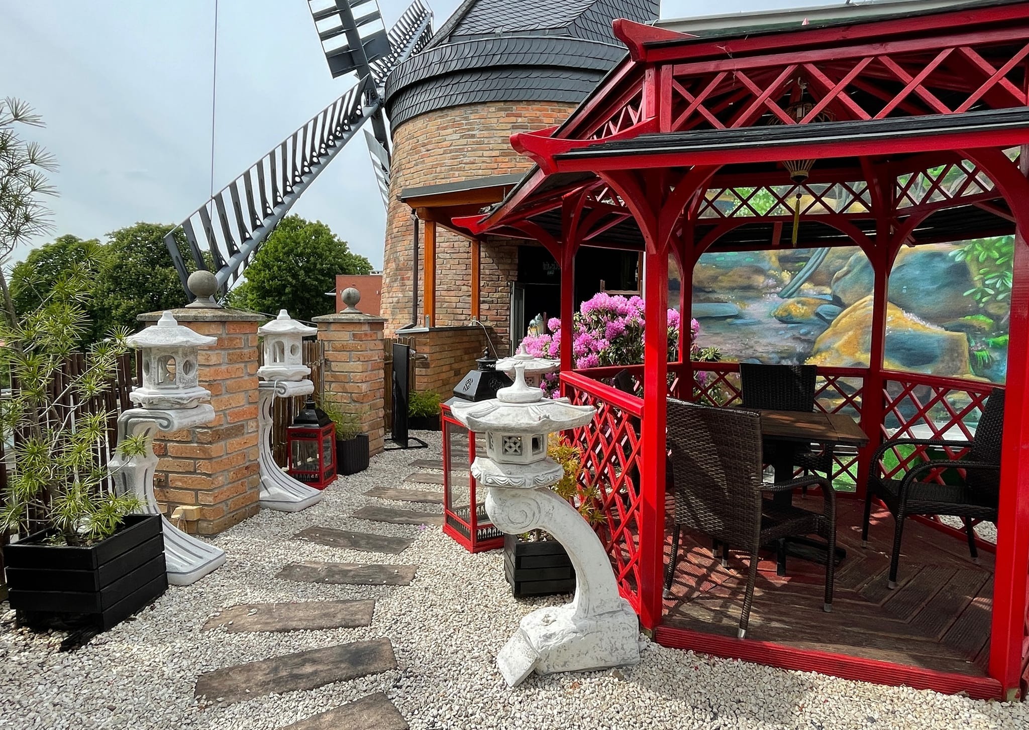 Ogród w stylu orientalnym z czerwonym pawilonem, kamiennymi latarniami, żwirową ścieżką i wiatrakiem w tle.