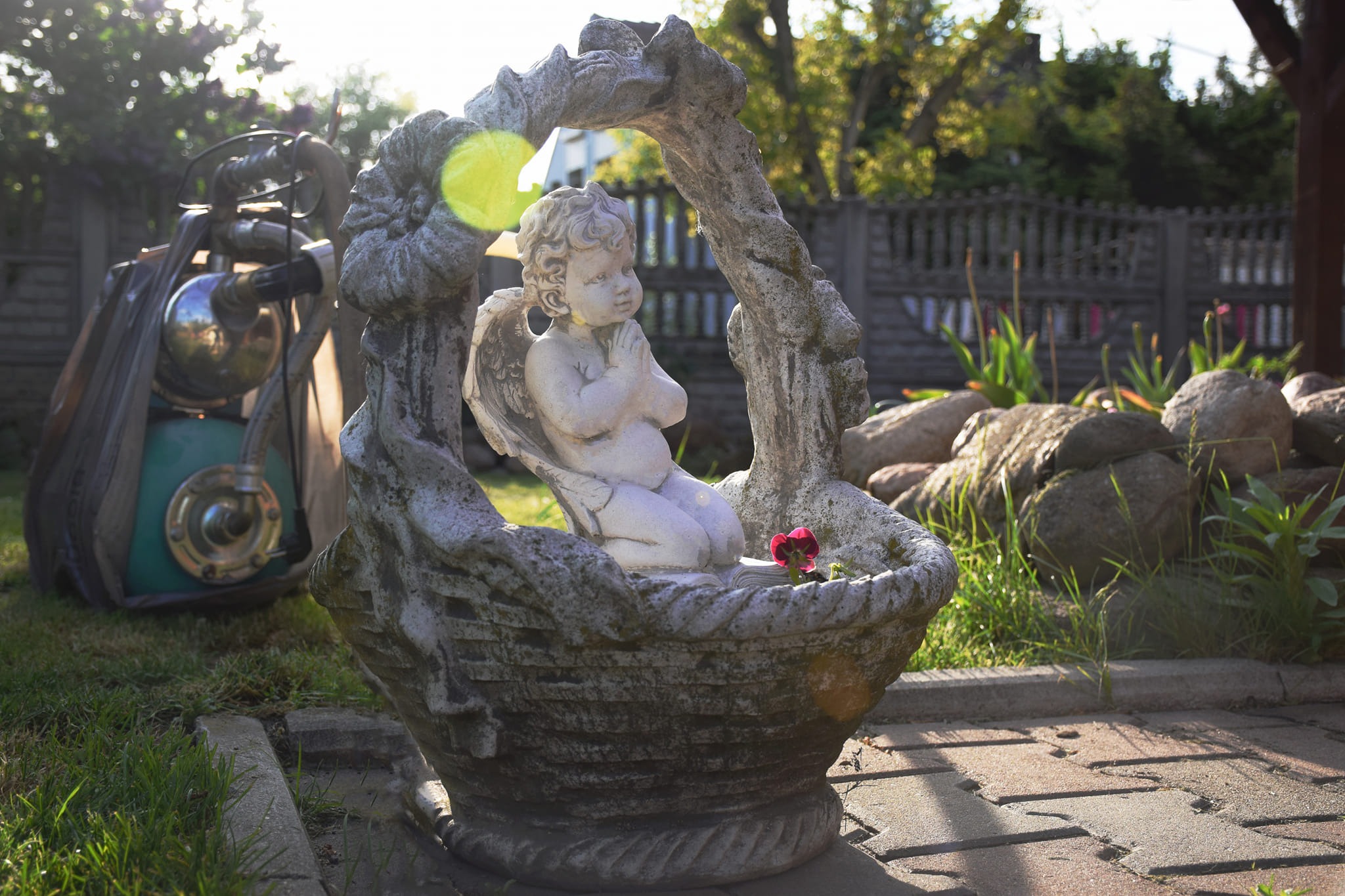 Ozdobna, kamienna donica w kształcie koszyka z figurką aniołka klęczącego w środku, z pojedynczym, czerwonym kwiatem, na tle ogrodu z elementami dekoracyjnymi i nietypowym, metalowym obiektem...