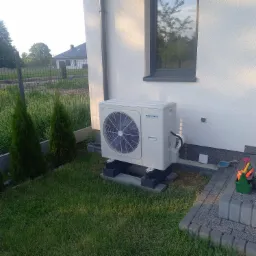 Biała, zewnętrzna jednostka klimatyzacyjna zamontowana na betonowych blokach przy ścianie domu z widocznym oknem i zielonym trawnikiem, obok narzędzia w zielonym koszyku.