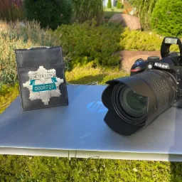 Odznaka detektywa z numerem 0008727 leżąca obok aparatu Nikon D3200 na laptopie HP Pavilion, w tle trawa i krzewy.