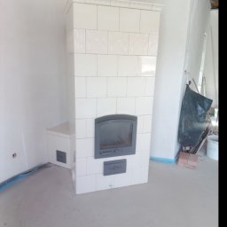 Piotr Mazurek - Biały piec kaflowy z szarymi drzwiczkami w surowym wnętrzu, widoczne ślady prac wykończeniowych na podłodze i ścianach.
