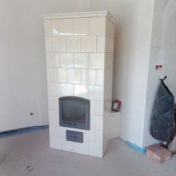 Piotr Mazurek - Biały, kaflowy piec z szarymi drzwiczkami w surowym wnętrzu, widoczne ślady po pracach wykończeniowych na ścianach i podłodze, zabezpieczenia folią w narożnikach.