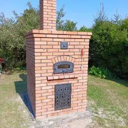 Nowo wybudowany, ceglany piec zewnętrzny z metalowymi drzwiczkami i kominem na tle zieleni i błękitnego nieba.