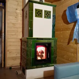 Stojący, zielony piec kaflowy z palącym się wewnątrz ogniem, umieszczony w rogu drewnianego pomieszczenia z boazerią. Widoczny fragment sofy.