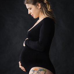 Aneta Wigłasz - Kobieta w czarnym body, profil, dłoń na ciążowym brzuchu, tatuaż mapy świata na udzie, ciemne tło.