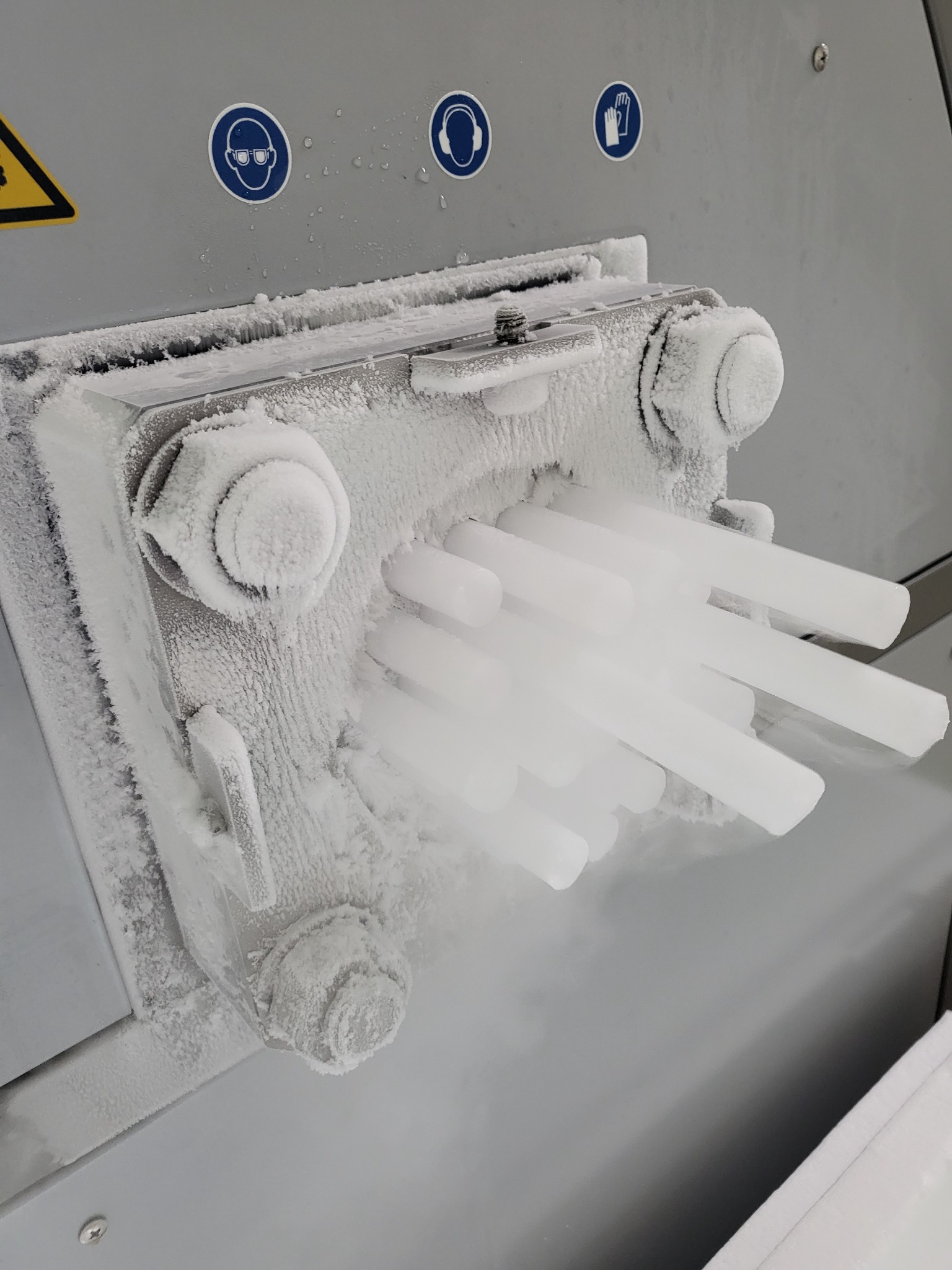 Zamrożone pręty w urządzeniu chłodniczym, pokryte szronem, z widocznymi znakami ostrzegawczymi dotyczącymi ochrony oczu, słuchu i rąk.