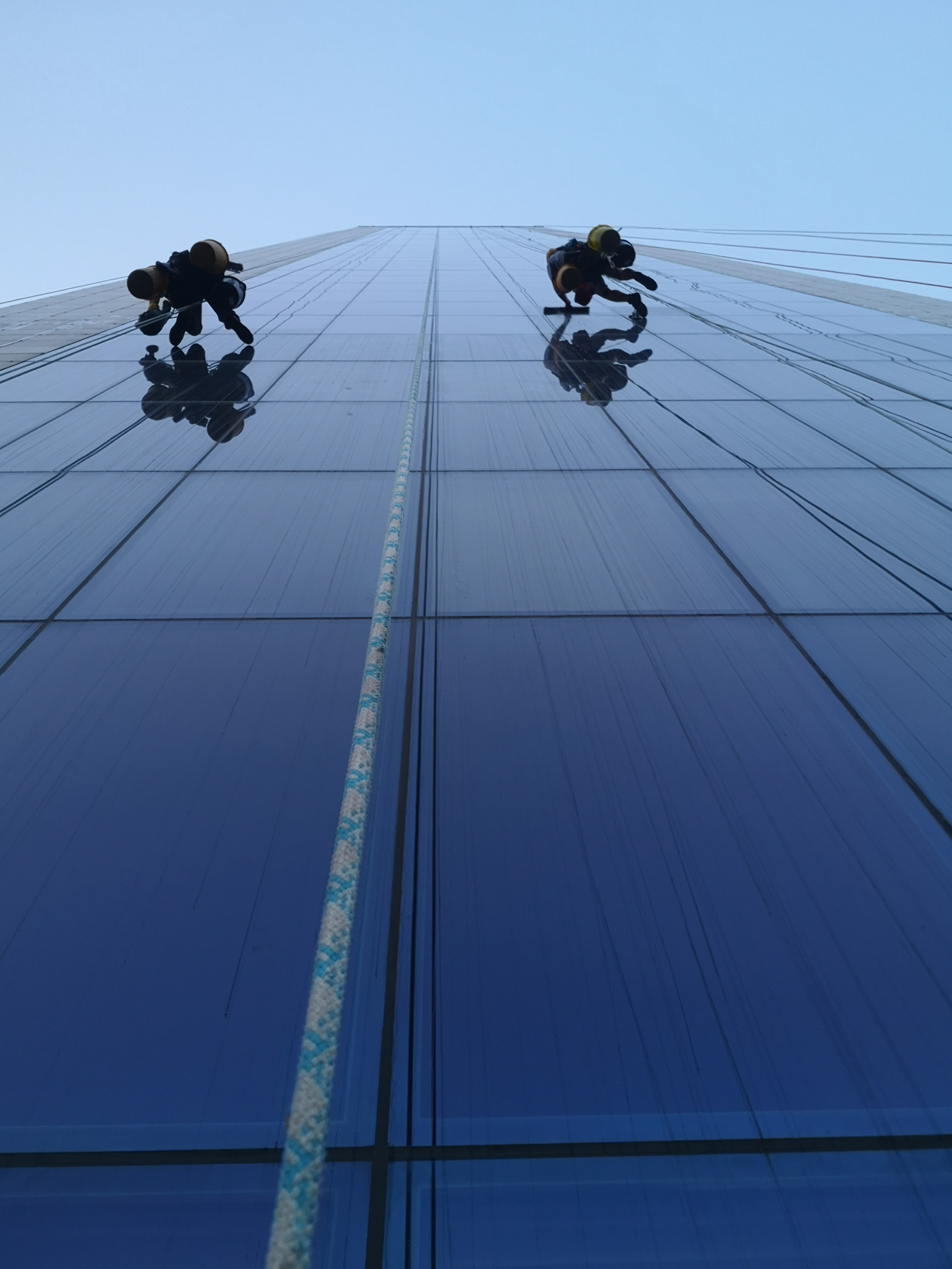 Dwóch alpinistów przemysłowych, zabezpieczonych linami, myje szklaną fasadę wysokiego biurowca, widoczne smugi brudu na oknach przed umyciem.