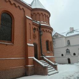 Zakład Budowlano Sztukatorski Włodarczak Krzysztof - Ceglany budynek z wieżyczką i oknem łukowym, pokryty śniegiem. Widoczny fragment schodów wejściowych i sąsiedni budynek w tle. Zimowy krajobraz.