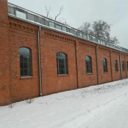 Zakład Budowlano Sztukatorski Włodarczak Krzysztof - Długa, czerwona ceglana ściana budynku z rzędami okien łukowych, pokryta śniegiem. Widoczne rynny i szklany dach. Zimowy krajobraz.