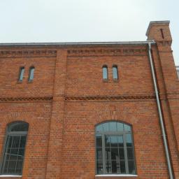 Zakład Budowlano Sztukatorski Włodarczak Krzysztof - Ceglana fasada budynku z łukowymi oknami i metalową rynną, widoczna w pochmurny dzień. Architektura z czerwonej cegły. Detale elewacji.