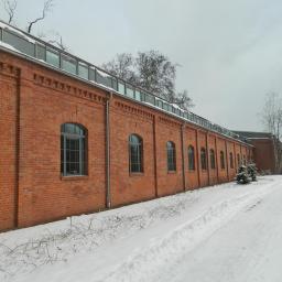 Zakład Budowlano Sztukatorski Włodarczak Krzysztof - Długi, ceglany budynek z rzędem okien łukowych, pokryty śniegiem. Widoczne ślady ośnieżonej drogi i drzewa bez liści. Zimowa sceneria.