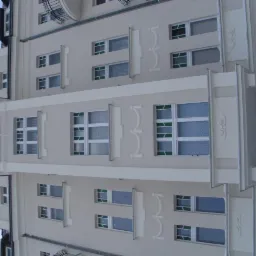 Odnowiona elewacja kamienicy z dekoracyjnymi detalami i białymi obramowaniami okien, widok z dołu na jasną fasadę budynku z elementami sztukaterii.