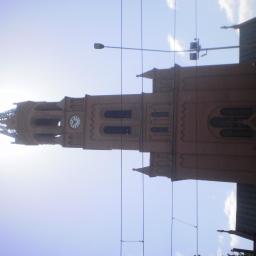 Zakład Budowlano Sztukatorski Włodarczak Krzysztof - Kościół z cegły z wieżą zegarową, widoczny z dołu pod kątem. Na pierwszym planie kable, w tle błękitne niebo i słońce. Nietypowe ujęcie.