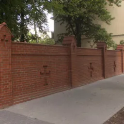 Ceglany mur z dekoracyjnymi elementami i krzyżami, oddzielający posesję. Widoczna brama wjazdowa i chodnik. Mur w odcieniach czerwieni, tło zielone drzewa i budynek.