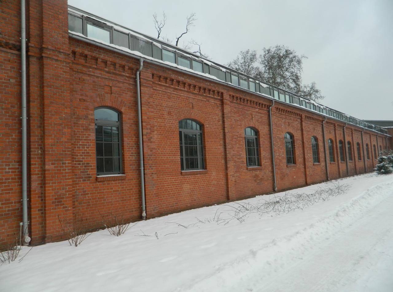 Długa, czerwona ceglana ściana budynku z rzędami okien łukowych, pokryta śniegiem. Widoczne rynny i szklany dach. Zimowy krajobraz.