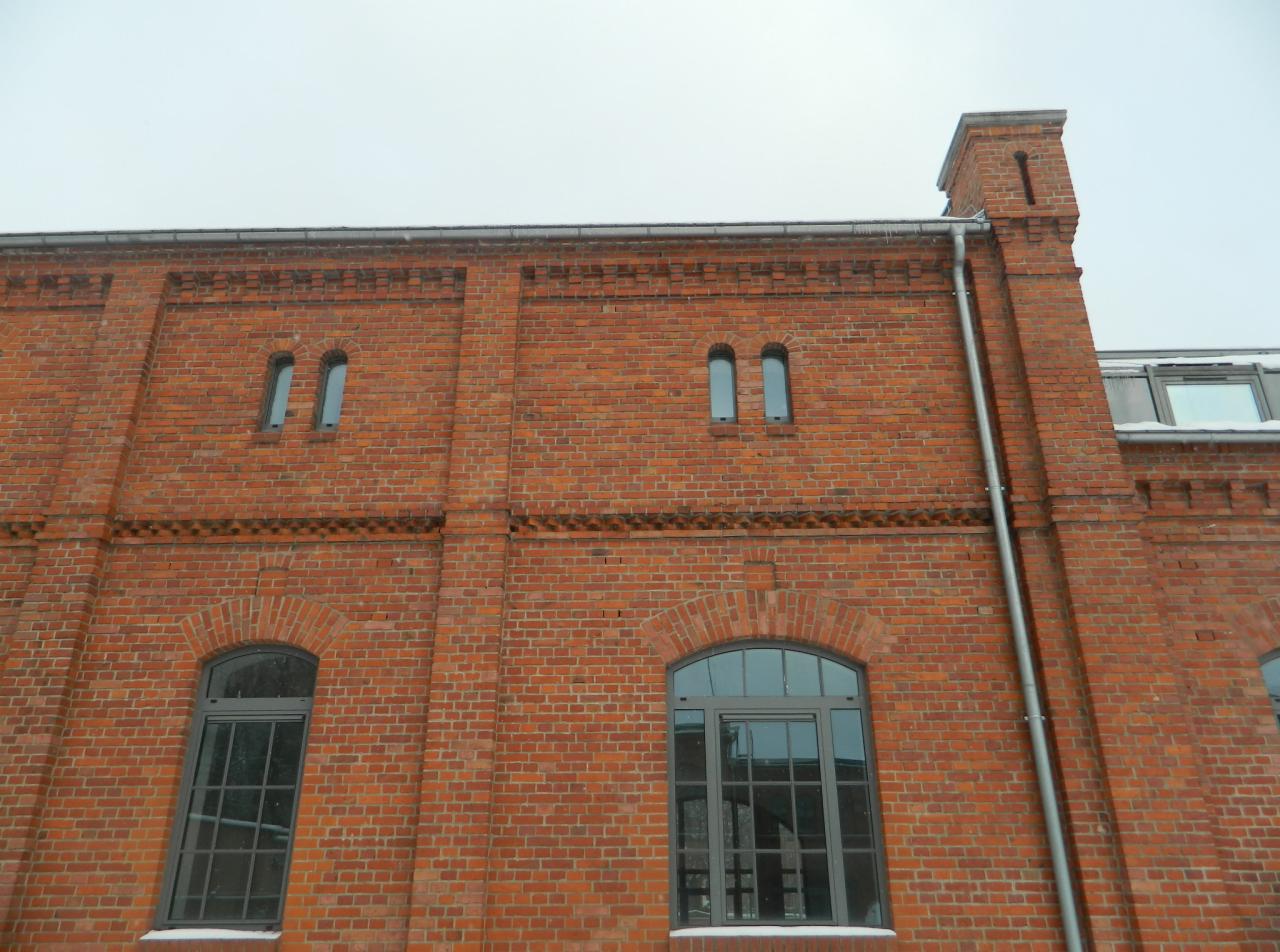 Ceglana fasada budynku z łukowymi oknami i metalową rynną, widoczna w pochmurny dzień. Architektura z czerwonej cegły. Detale elewacji.