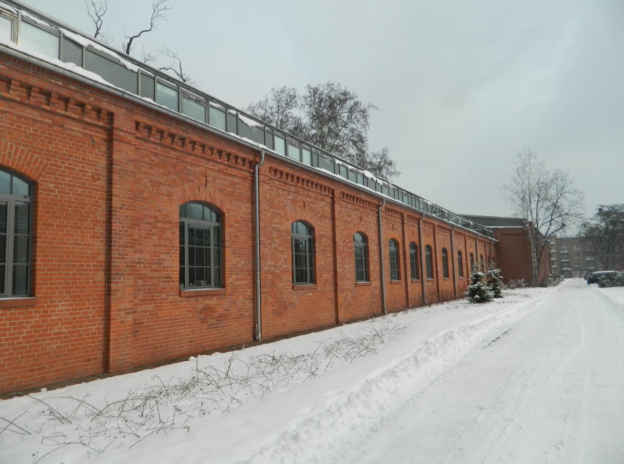 Długi, ceglany budynek z rzędem okien łukowych, pokryty śniegiem. Widoczne ślady ośnieżonej drogi i drzewa bez liści. Zimowa sceneria.