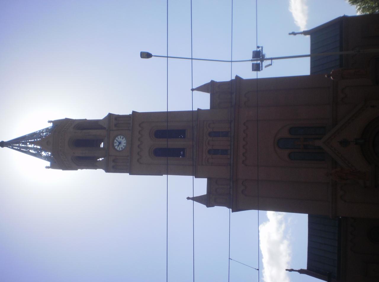 Wieża kościelna z zegarem w słoneczny dzień, widok z dołu. Na pierwszym planie kable elektryczne. Budowla z cegły, detale architektoniczne.