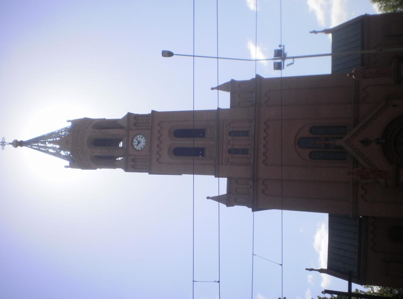 Kościół z cegły z wieżą zegarową, widoczny z dołu pod kątem. Na pierwszym planie kable, w tle błękitne niebo i słońce. Nietypowe ujęcie.