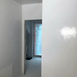 Świeżo pomalowane na biało wnętrze z widocznymi śladami po natryskowym malowaniu, zabezpieczone folią okno w tle.