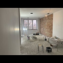 Dez-bud - Wnętrze w trakcie prac remontowych we Włocławku: Częściowo pomalowane ściany, surowa cegła, sprzęt malarski, drabina i materiały budowlane.