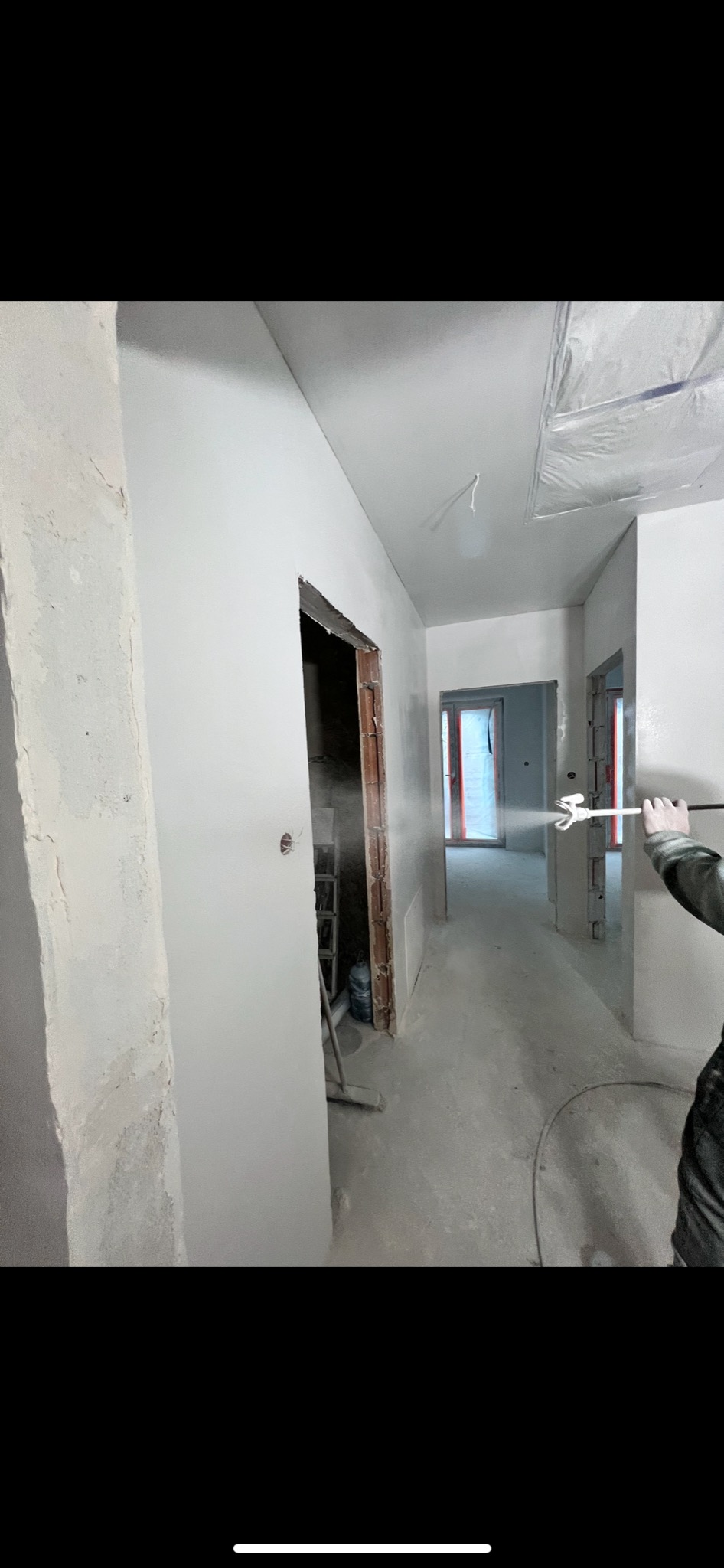 Malowanie natryskowe ścian w korytarzu, widoczne ślady farby w powietrzu, surowe wykończenie ościeżnic, przygotowanie do dalszych prac wykończeniowych.